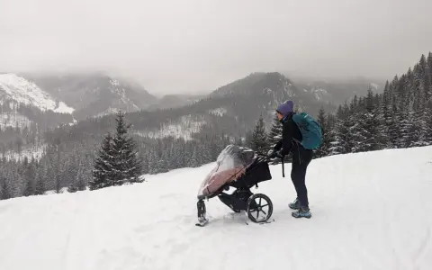 Zimowe Tatry z wózkiem. Kobieta w czapce i z plecakiem pcha wózek po ośnieżonym stoku, podziwiając górski krajobraz.