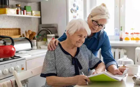 Dos mujeres mayores revisan documentos en la cocina. Una apoya a la otra, quizás tramitando la pensión de orfandad mayores de 30 años.