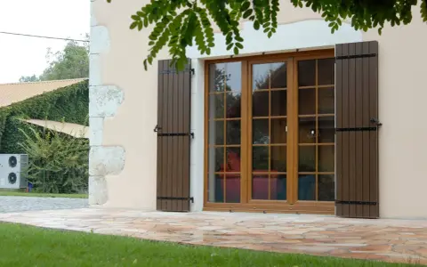 Fenêtre en bois avec volets bruns, matériaux menuiserie de qualité. Le jardin verdoyant et la façade en pierre complètent le tableau.