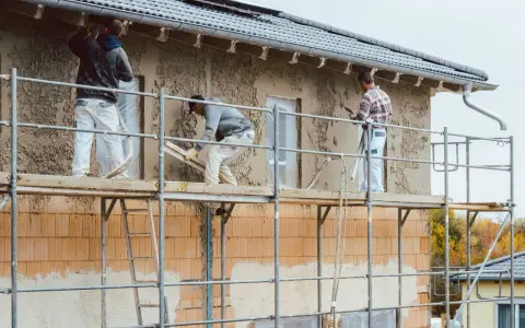 Pracownicy na rusztowaniu wykonują tynkowanie elewacji domu, nakładając zaprawę na ściany.