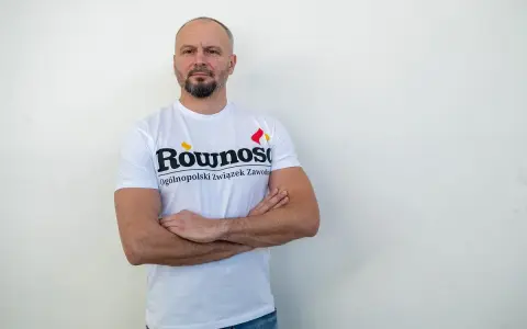 Mężczyzna w koszulce z napisem "Równość Ogólnopolski związek zawodowy" stoi z rękami skrzyżowanymi na piersi.