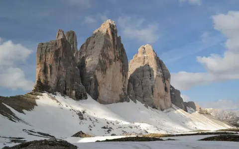 Majestatyczne **Tre Cime** w Dolomitach, ośnieżone zbocza i błękitne niebo z chmurami.