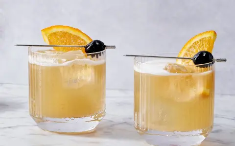 Dwa drinki Rusty Rat z pomarańczą i wiśnią na patyczku, podane w szklankach z lodem.