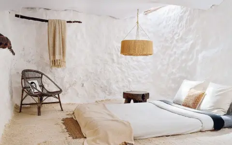 Habitación rústica con paredes de encalar, cama baja, silla de mimbre y manta de lana.