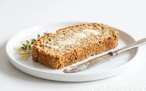 Keto chleb z mąki migdałowej i kokosowej, posmarowany masłem, na talerzu z płatkami migdałów i gałązką tymianku. Obok leży nóż.