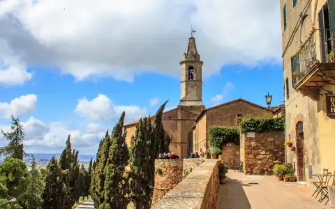 Un borgo nei comuni Val d'Orcia: un campanile svetta tra cipressi e case antiche, con un bar all'aperto.