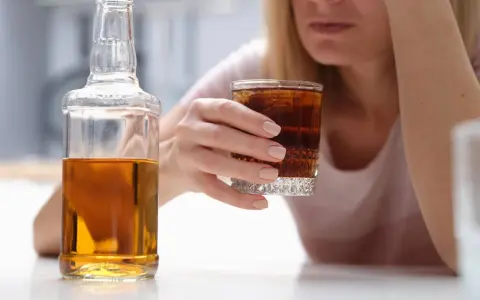 Kobieta z kieliszkiem drinka i butelką alkoholu. W tle widać jej twarz, która wyraża smutek. To może być wszywka alkoholowa.