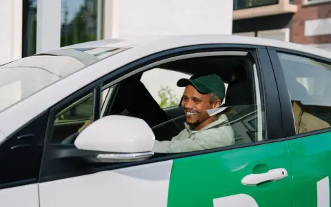 Lächelnder Mann mit grüner Kappe am Steuer eines weißen Autos mit grünen Bolt-Aufklebern. Er wird bald ein Bolt-Fahrer sein.