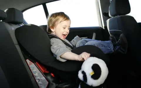Ein fröhliches Kind sitzt im Kindersitz im Auto und spielt mit einem Panda-Stofftier.