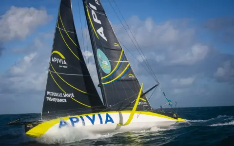 Le voilier Apivia, skippé par Alex Thomson, fend les vagues sous un ciel bleu et nuageux.