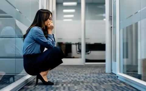 Frau kauert im Büroflur, Hände vor dem Mund, die Augen geschlossen. Sie scheint von **Angst vor der Arbeit** überwältigt zu sein.