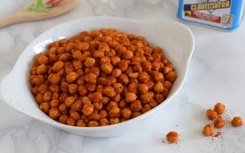 Un tazón blanco lleno de garbanzos fritos crujientes y especiados. Una cuchara de madera decorada y una lata de especias están al fondo.