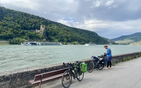 Mann packt Gepäck für eine Fahrradtour am Rhein. Im Hintergrund Burg und Fluss.