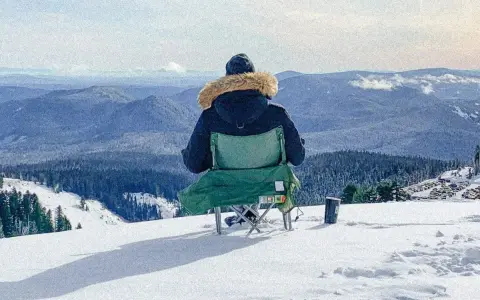 Persona sentada en silla plegable en la nieve, mirando un paisaje montañoso cubierto de árboles.