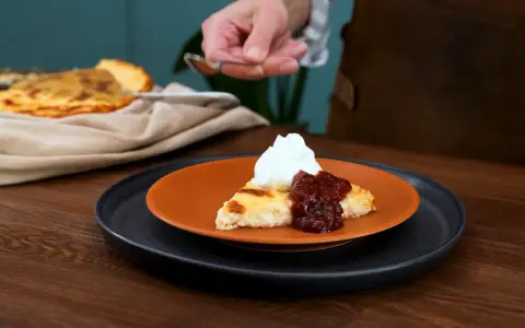 En bit krämig ostkaka med keso, toppad med vispgrädde och sylt. Serveras på en orange tallrik.