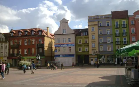 Rynek w Chojnicach z kolorowymi kamienicami, w tym ratusz. Ludzie spacerują po brukowanym placu.