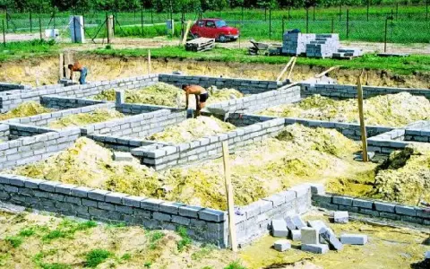 Budowa fundamentu z bloczków betonowych. Dwóch robotników pracuje przy wznoszeniu ścian fundamentowych.