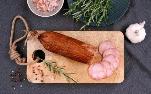 Kiełbasa żywiecka na desce, obok gałązka rozmarynu, czosnek i kolorowy pieprz. Kilka plastrów już pokrojonych.