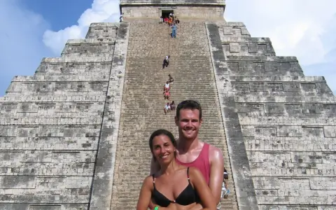 Coppia sorridente davanti alla piramide di Kukulkan, con turisti che salgono le scale del chichen itza interno.