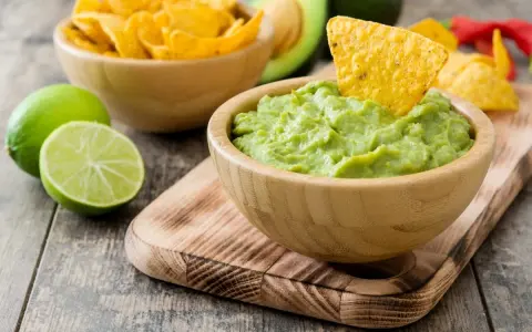 Pyszne guacamole w drewnianej misce, idealne do chrupania z nachos. Obok limonki i miska pełna chipsów.