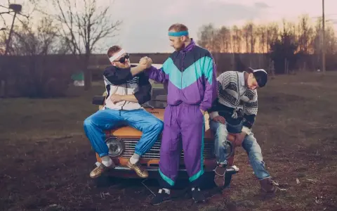 Trzech kumpli w dresach z lat 90. szykuje się na imprezę. Jeden siedzi na masce, drugi podaje mu rękę, trzeci odpoczywa obok.