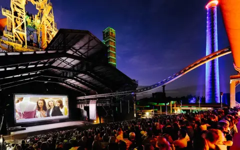 Ein belebtes **freiluft kino** mit vielen Zuschauern, die einen Film auf einer großen Leinwand vor einer beeindruckenden Industriekulisse mit bunten Türmen und Stahlkonstruktionen genießen.