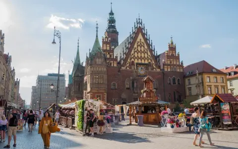 Tłum ludzi spaceruje po Rynku we Wrocławiu, podziwiając zabytkowy ratusz i letnie stoiska.