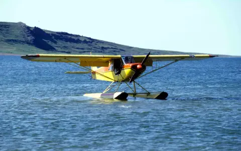 Gelbe Piper Super Cub mit Schwimmern auf ruhigem Wasser, im Hintergrund eine grüne Hügelkette.