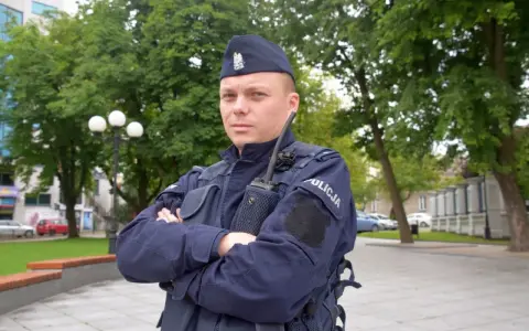 Policjant w granatowym mundurze, w sprawie umundurowania policjantów, stoi na tle zieleni.