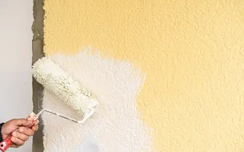 Hand streicht mit einer Farbrolle über eine raue, gelbe Wand. Der Putz wird gerade weiß gestrichen.