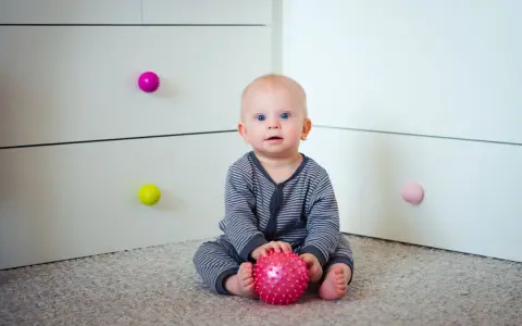 7 miesięczne dziecko z niebieskimi oczami siedzi na dywanie, trzymając w rączkach różową piłeczkę.