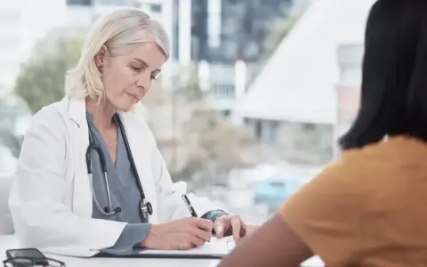Lekarka w białym kitlu z receptą w ręku, pacjentka w żółtej bluzce. Wizyta u lekarza, może l4 na jeden dzień.