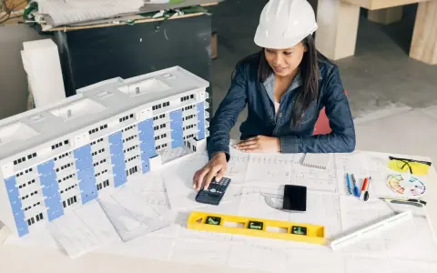 Architektka w kasku analizuje projekt budynku, używając kalkulatora. To może być związane z bms co to jest za system.