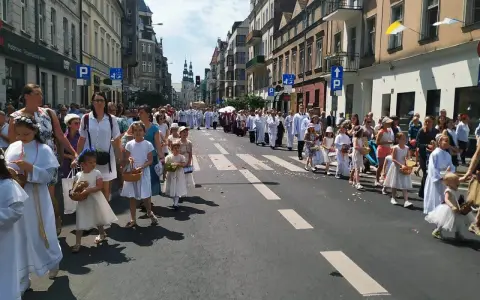 Procesja dzieci w białych strojach, niosących koszyki, idzie ulicą. W tle widać budynki i ludzi, co może symbolizować sekularyzację społeczeństwa.