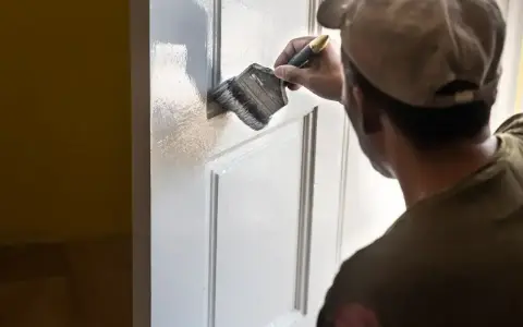 Mężczyzna w czapce z daszkiem maluje białe drzwi pędzlem. Odświeża wnętrze domu.