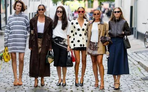 Sechs Frauen in stylischen Outfits, darunter ein gestreiftes Kleid und ein Blazer mit nordischen Mustern, posieren auf einer Kopfsteinpflasterstraße.