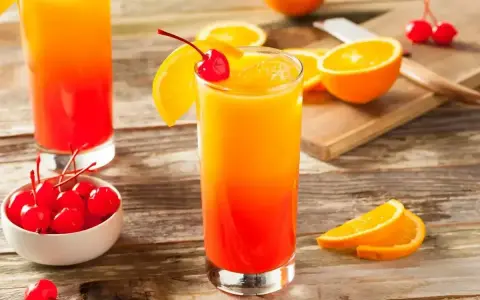 Dwa drinki z whisky i sokiem pomarańczowym, ozdobione wiśniami i plasterkami pomarańczy, na drewnianym stole.