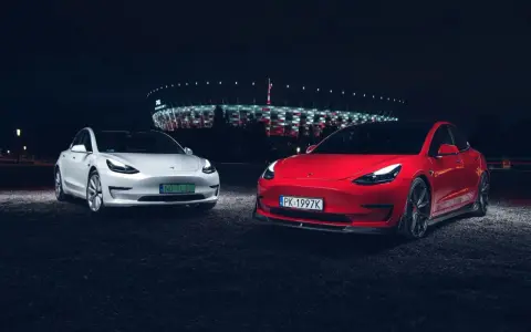 Dwa modele Tesla, biały i czerwony, zaparkowane nocą przed oświetlonym stadionem.
