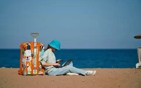Dziewczyna w niebieskim kapeluszu siedzi na plaży z walizką, laptopem i telefonem, planując, jak tanio podróżować.