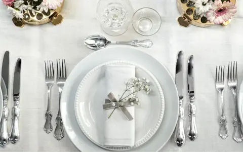 Ett elegant dukat bord med vita tallrikar, servetter dekorerade med blommor och vackra, silverfärgade engångsbestick.