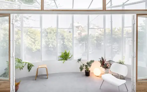 Interno luminoso con piante, sedia bianca e sgabello in legno, affacciato su una strada alberata da una veranda balcone.