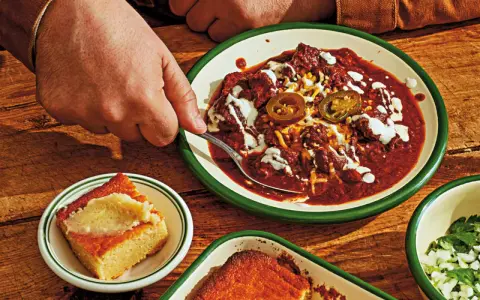 En hand tar en sked ur en skål med mustig texas chili, toppad med jalapeños och gräddfil. Bredvid ligger gyllene majsbröd.