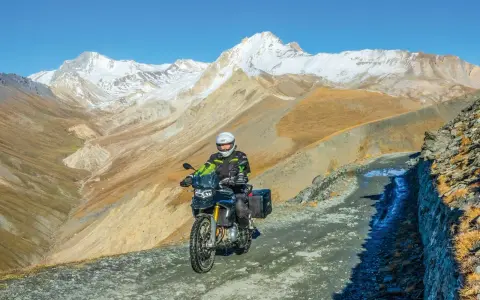 Un motociclista avventuroso affronta un sentiero impervio con la sua jafferau moto, circondato da maestose montagne innevate e paesaggi aridi.
