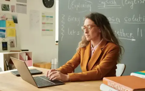 Nauczyciel początkujący w okularach pracuje przy laptopie w klasie. Na tablicy napisy po angielsku.