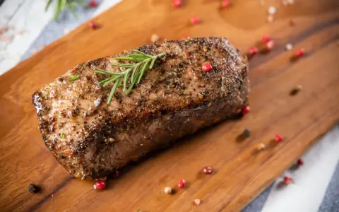 Un délicieux roti de boeuf, assaisonné et garni de romarin, repose sur une planche en bois, entouré de grains de poivre rose et noir.