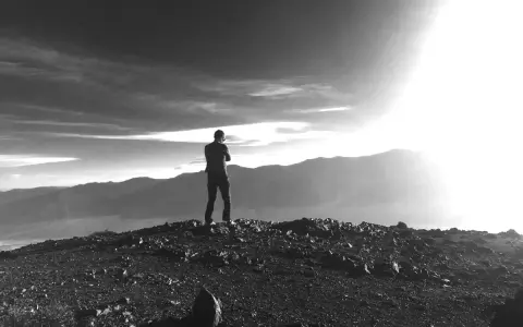 Ein Mann steht auf einem felsigen Gipfel und blickt in die Ferne. Die Sonne bricht durch die Wolken und beleuchtet die Berge. Vielleicht fragt er sich, was ist Weisheit.