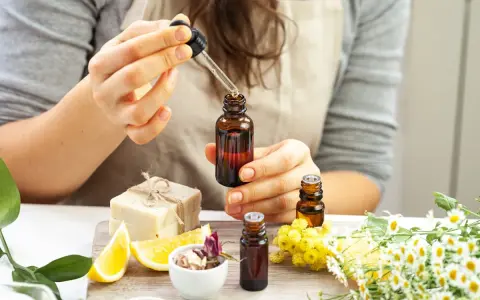 Une personne prépare un parfum huile essentielle, ajoutant une goutte d'ambre dans un flacon. Des fleurs, des citrons et du savon sont disposés autour.