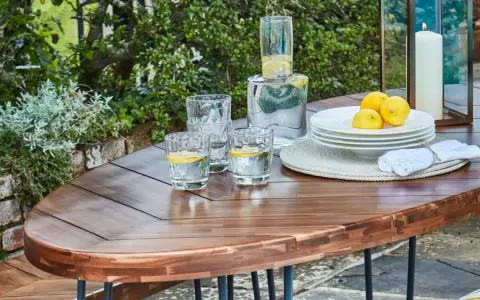 Table en bois couleur acacia, prête pour un apéritif en plein air avec des verres d'eau citronnée, des citrons et une bougie.