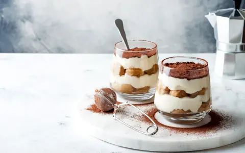 Dwa pucharki z kremowym tiramisu bez jajek, posypane kakao. Obok sitko do kakao i kawiarka.