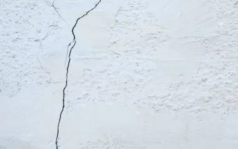 Un muro de casa blanco con una grieta oscura que lo atraviesa. La textura es rugosa y desigual.